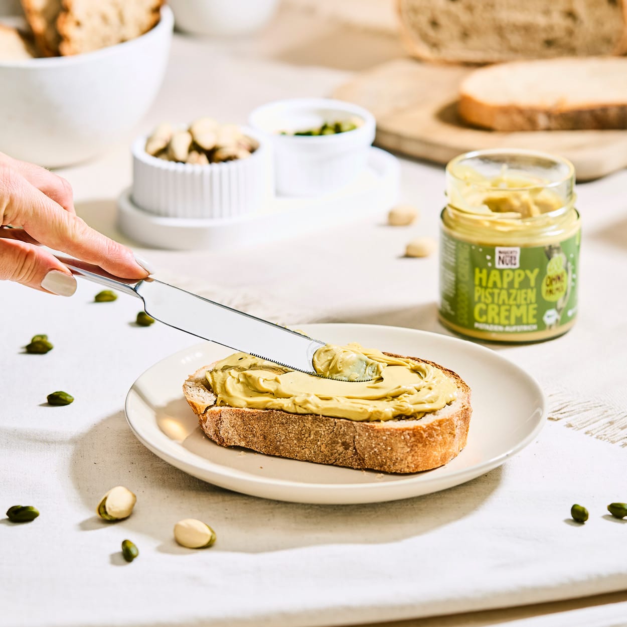 Happy Pistazien Creme auf Brot gestrichen mit Messer und Glas im Hintergrund