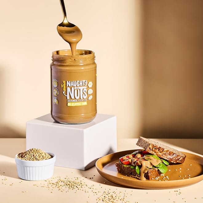 Löffel taucht in ein Glas Bio Sesammus Tahin dunkel, daneben Brot mit Aufstrich und Sesamsamen